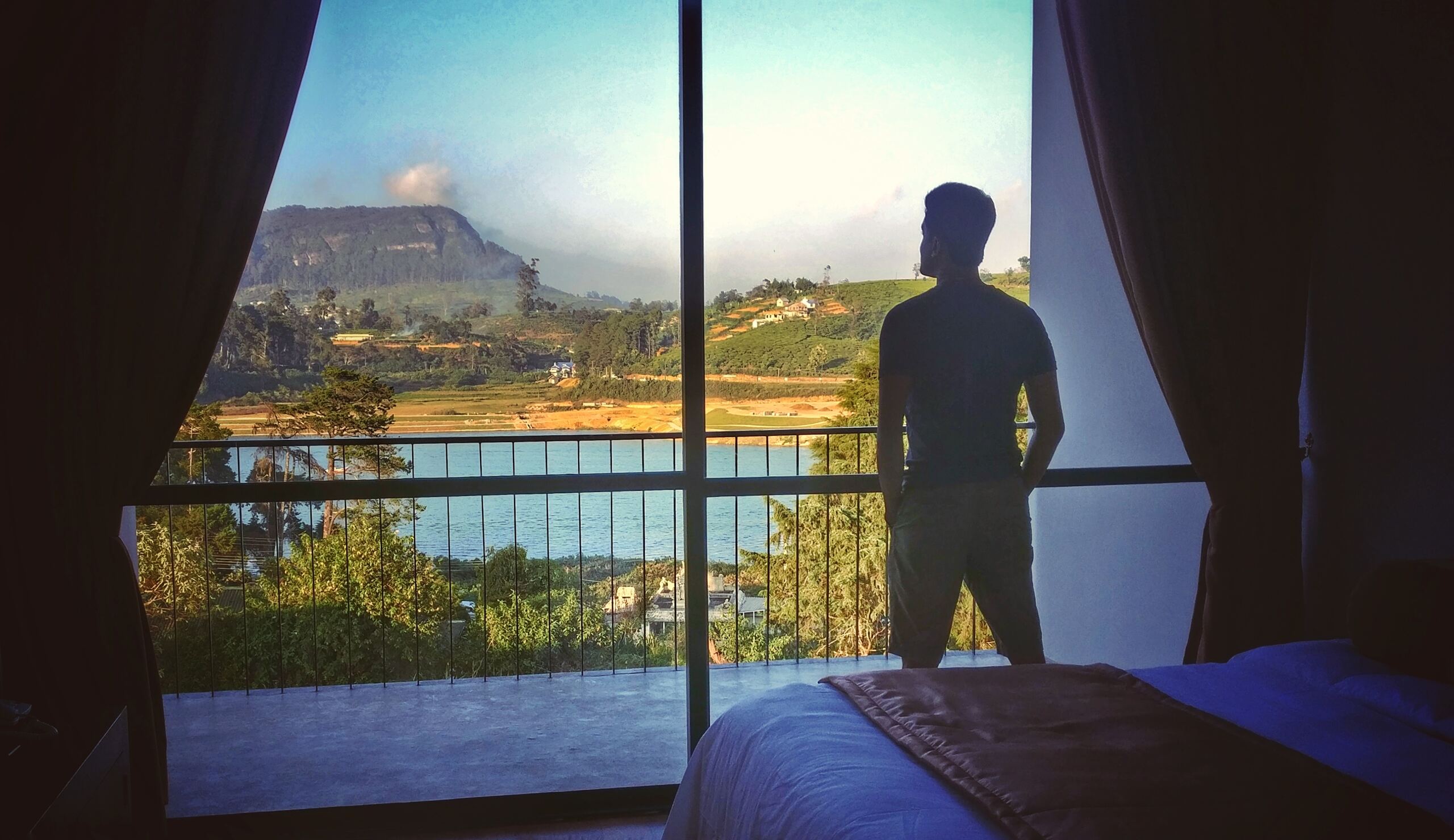 Hirantha Perera silhouetted at a floor-to-ceiling window, looking out over a lake and the hills of Sri Lanka